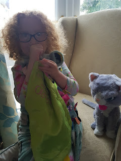 An image of a four year old white girl with curly red hair, wearing purple glasses, sucking her thumb and holding a green Taggie and a blue one. A toy cat sits next to her.
